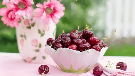 Cherries in a pink flower-shaped bowl near a pink floral vase containing pink flowers.