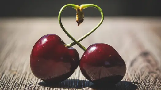 Two cherries looped at the stem, sitting on a wooden surface outdoors.