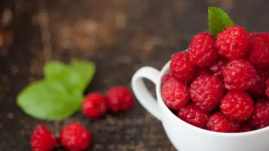 A white teacup full of raspberries, with leaves in the background.