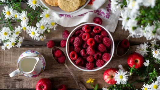 A white bowl full of raspberries, decorated with nearby daisies, a bowl of biscuits, apples, spoons, and a pitcher decorated with flowers.