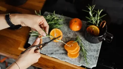 A person cutting herbs over orange slices with a cup of herbs, cinnamon, and an orange slice on the side.