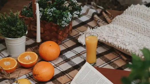 A glass of orange juice, near some oranges, plants, and woven cloth.