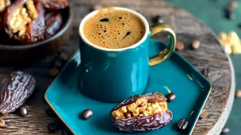 A stuffed date and a blue mug of coffee sitting on a blue platter, with a wooden bowl full of stuffed dates out of focus.