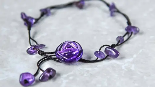 A smoothed purple amethyst shaped into a small rose on a black braided cord adorned with small smoothed purple amethyst drops resting on a marbled white surface.