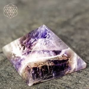 A smoothed purple amethyst pyramid with clear and white inclusions resting atop a coarse grey surface. Hyperlinks to Conscious Items product "The Pyramid of Devotion" in a new tab.