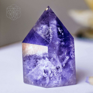 A purple faceted Amethyst shard with cloudy white coloration infusing the purple tones, resting on a white surface. Hyperlinks to Conscious Items Product "The Artist's Sleep Stone" in a new tab.