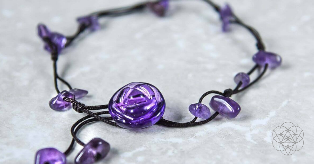A smoothed purple amethyst shaped into a small rose on a black braided cord adorned with small smoothed purple amethyst drops resting on a marbled white surface. Hyperlinks to Conscious Items product "The Emotional Healing Anklet" in a new tab.