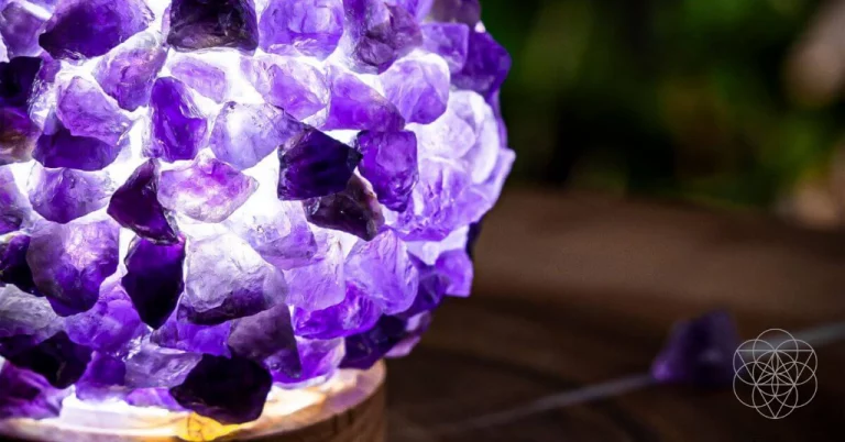 A purple amethyst cluster hollowed and lit from the interior with white light, set atop a circular wood base. Hyperlinks to Conscious Items product "The Intuition Lamp" in a new tab.