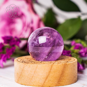 A smoothed purple Amethyst crystal ball resting atop a circular wooden base in front of bright pink carnations. Hyperlinks to Conscious Items product "The Stress Relief Lamp" in a new tab.