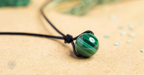 A smooth shiny Malachite bead with alternating bands of varying shades of green strung on a black braided cord resting on a tan surface near plant grains and seeds. Hyperlinks to Conscious Items product "The Detoxification Necklace" in a new tab.