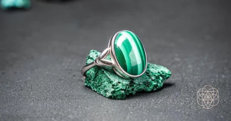 A green malachite cabochon ring with vertical line coloration starting lightest green in the center line and alternating dark and light outward, resting atop a raw malachite shard. HyperLinks to Conscious Items product "The Heart Healing Ring".