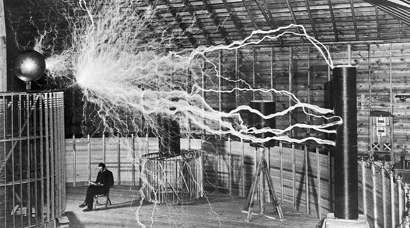 Nikola Tesla sitting with his equipment. A stream of electrical energy travels between the equipment.