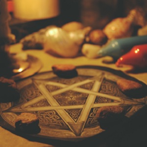 A pentacle lying flat with five stones laying atop it. A red and a blue candle are in the background. Witchcraft is a theory of magic and a discipline that teaches you how to harness and direct universal energies. • Beginner Witch Essentials