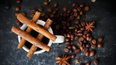 A bird-eye view of cinnamon sticks in a hashtag pattern on top of a cup of coffee, near coffee beans and anise seeds.