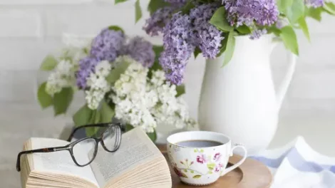 Two vases of purple and white lilac flowers, near a book with glasses on it and a teacup of coffee.