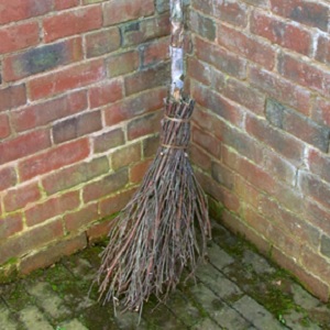 A besom leaning against a red brick wall on a mossy floor.