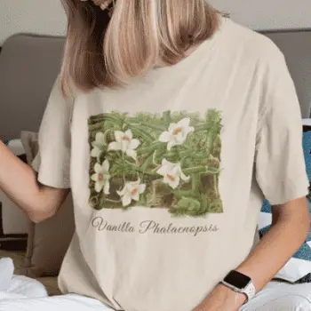 A woman wearing a soft cream t-shirt with a vanilla orchid graphic. Text on the bottom of the vanilla orchid graphic says: "Vanilla Phalaenopsis."