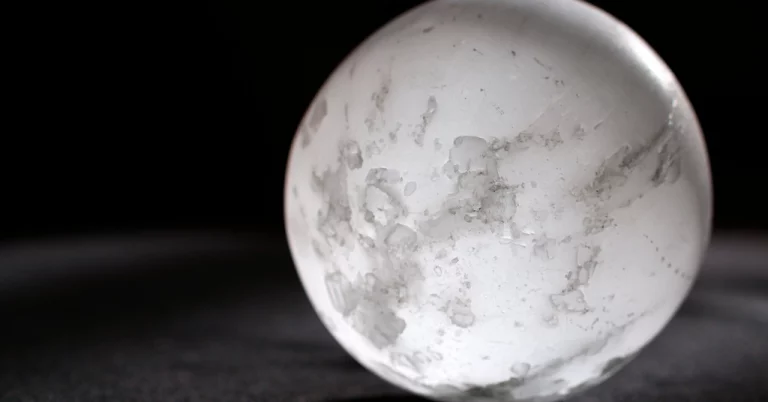 A selenite crystal sphere that almost resembles the moon with a subtle glow, sitting on a darkened surface against a dark background.