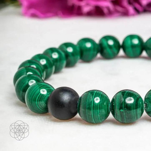 A bracelet of smoothed malachite beads with a banded pattern of varying shades of green with a single smoothed onyx bead, resting on a white foreground with a bright pink carnation in the background. Hyperlinks to Conscious Items Product "The Anti-Anxiety Double Bracelet Set" in a new tab. | Malachite Metaphysical Properties and Meaning |