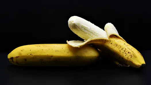 Two slightly ripened bananas, one peeled and one unpeeled, on a black surface.