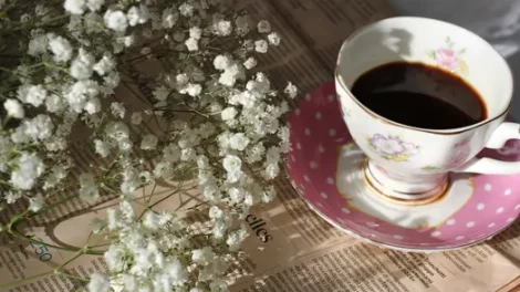 Coffee in a teacup near baby's breath flowers on a newspaper.