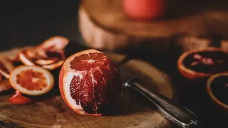 A blood orange with part of its peels cut off sitting on a circular slab of wood next to a weathered metal knife.