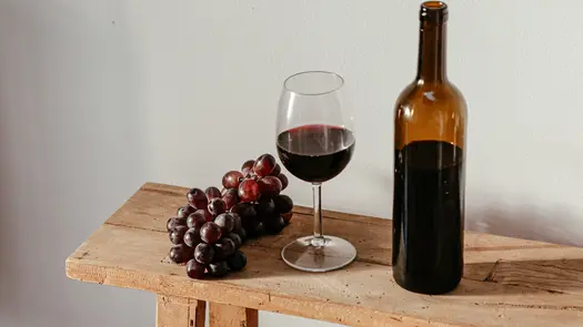 Grapes near a glass of wine and a wine bottle on a wooden shelf.