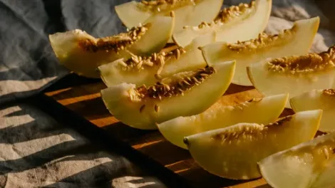 Slices of honeymelon on a wooden plank, resting on a gray cloth with a warm light shining through the honeymelon slices.