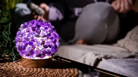 A lit amethyst-encrusted lamp sitting on a woven mat, with a person sitting cross-legged in the background holding a sage smudging stick.