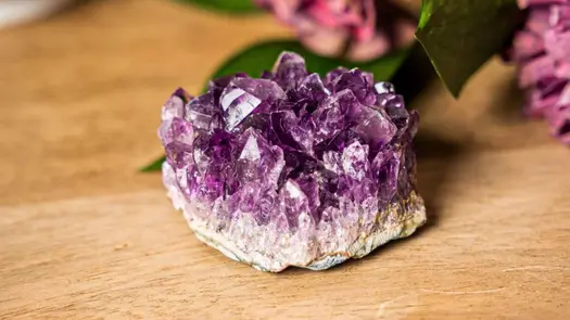 An amethyst cluster from Conscious Items resting on a wooden table near purple flowers.