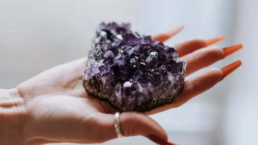 A person with long, manicured nails holding an amethyst cluster in the palm of their hand.