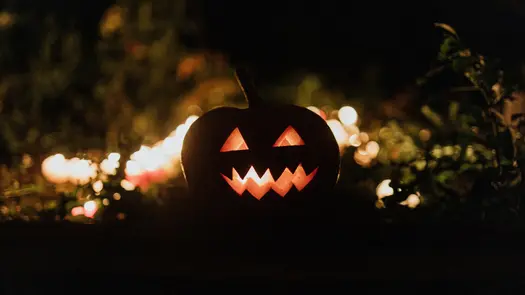 A jack o' lantern sitting in grass outside at night lit by lights in the background.