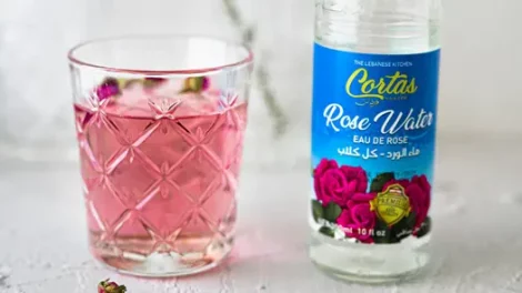 A glass cup full of rose water next to an empty rose water bottle.