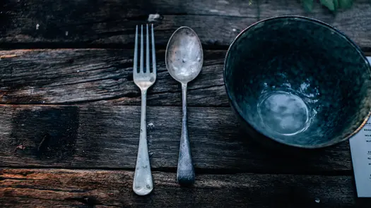 An empty dark teal bowl next to a worn fork and spoon sitting on a wooden table.
