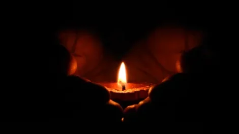A person holding a lit tealight candle in their hands in the dark.