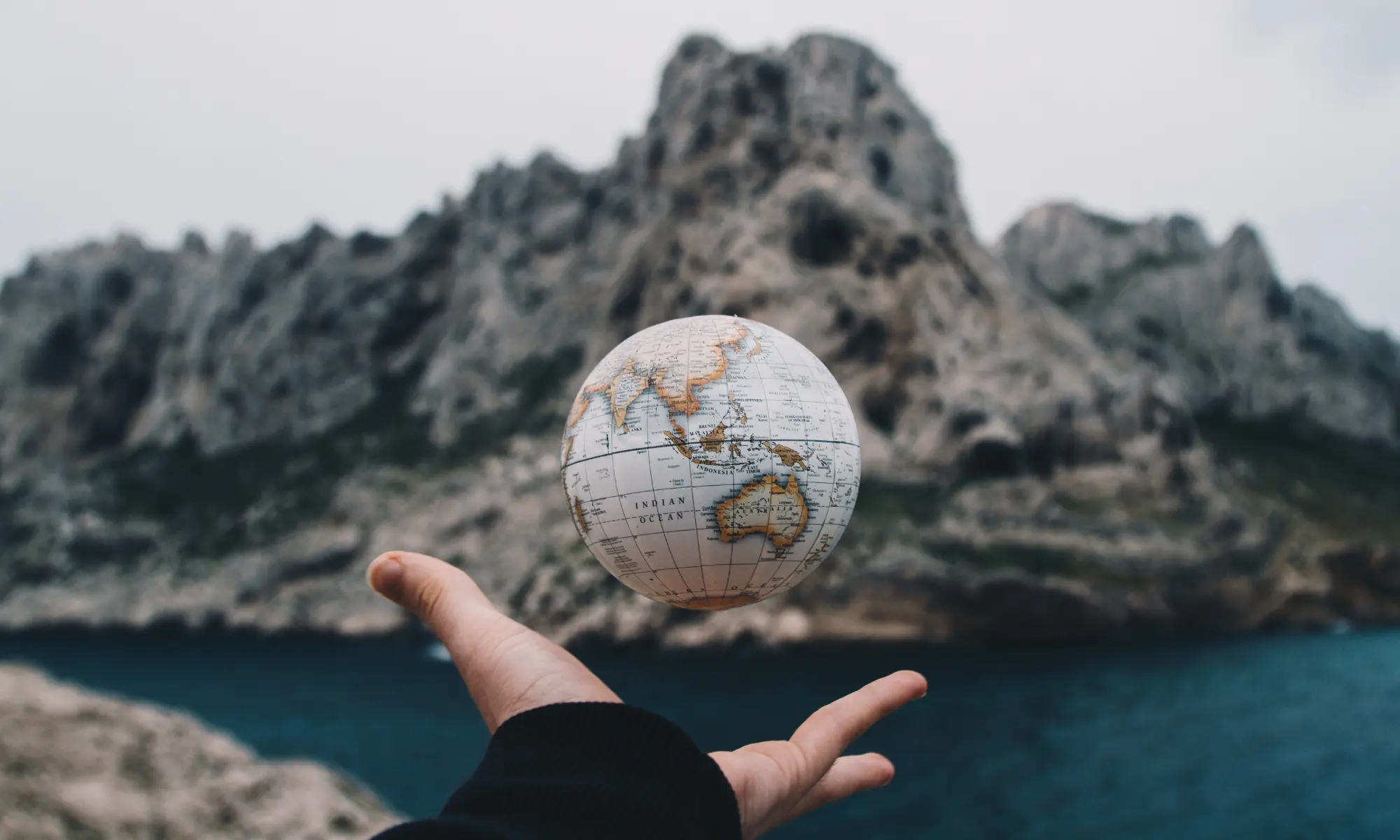 A person tossing a globe in the air with mountains and a lake in the background.