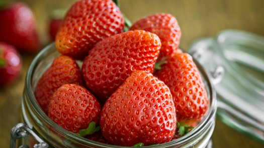 A glass jar filled with strawberries. The strawberries are a deep red color, with a few slightly lighter red strawberries mixed in. The leaves on the strawberries are a bright green color. The jar is a clear glass, and it has a metal lid with a white stripe.
